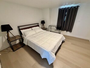 Ein ordentlich bezogenes Bett mit weißer Decke, zwei Nachttische mit Lampen, ein Fenster mit Vorhängen und ein Lichtschalter an der Wand.