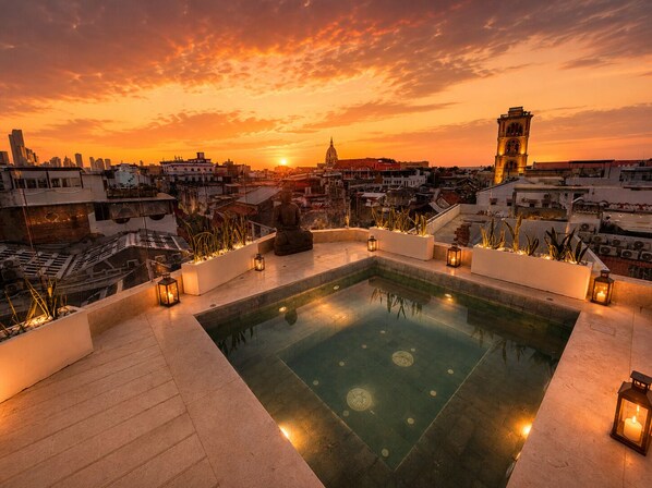 Ein Pool auf dem Dach mit einer Buddha-Statue, umgeben von Laternen, mit Blick auf eine Stadtlandschaft im Sonnenuntergang.