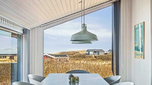 Et spiseområde med et bord og stole, en hængende pendellampe samt udsigt til et mark og bygninger gennem store vinduer.