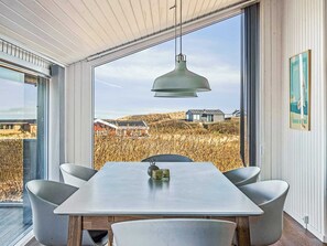 Ein Essbereich mit Tisch und Stühlen, einer hängenden Pendelleuchte sowie Blick auf ein Feld und Gebäude durch große Fenster.