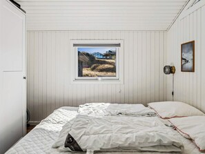 Un dormitorio con cama, ventana con vista a una casa y paisaje, lámpara de pared y un cuadro enmarco.