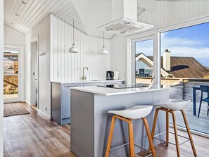 Eine moderne Küche mit einer großen Kochinsel, Pendelleuchten und Blick auf ein Haus durch ein großes Fenster.