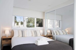 A hotel room with a large bed, two bedside tables with lamps, a large mirror, and windows with a view of trees and buildings. - Stylish 1BR Near CBD | WFH & Pet Friendly (Glebe)