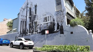 En bygning med et muralmaleri af historiske personer, en sporvogn, parkerede biler og en blå himmel.