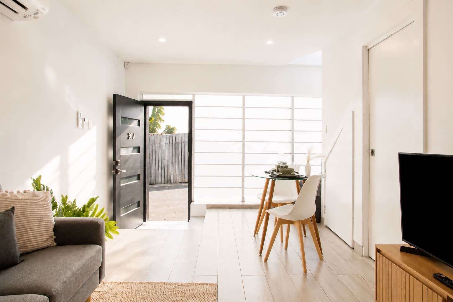 A modern living room with a sofa, a dining table with chairs, a television on a wooden stand, and a sliding glass door leading to an outdoor area.