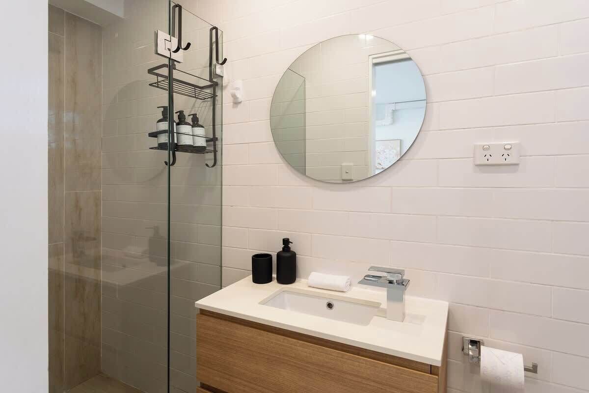 A modern bathroom with a glass shower enclosure, a round mirror, a white sink with a wooden vanity, and a towel rack.