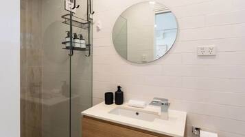 A modern bathroom with a glass shower enclosure, a round mirror, a white sink with a wooden vanity, and a towel rack.