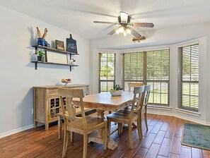 A dining area with a wooden table and chairs, a ceiling fan, and a window with blinds.