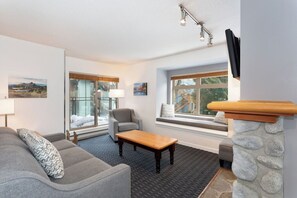 A living room with a sofa, armchair, and coffee table, large windows with views, and a stone fireplace. - Whistler Creekside Ski in/Ski out Condo with Pool (Whistler)