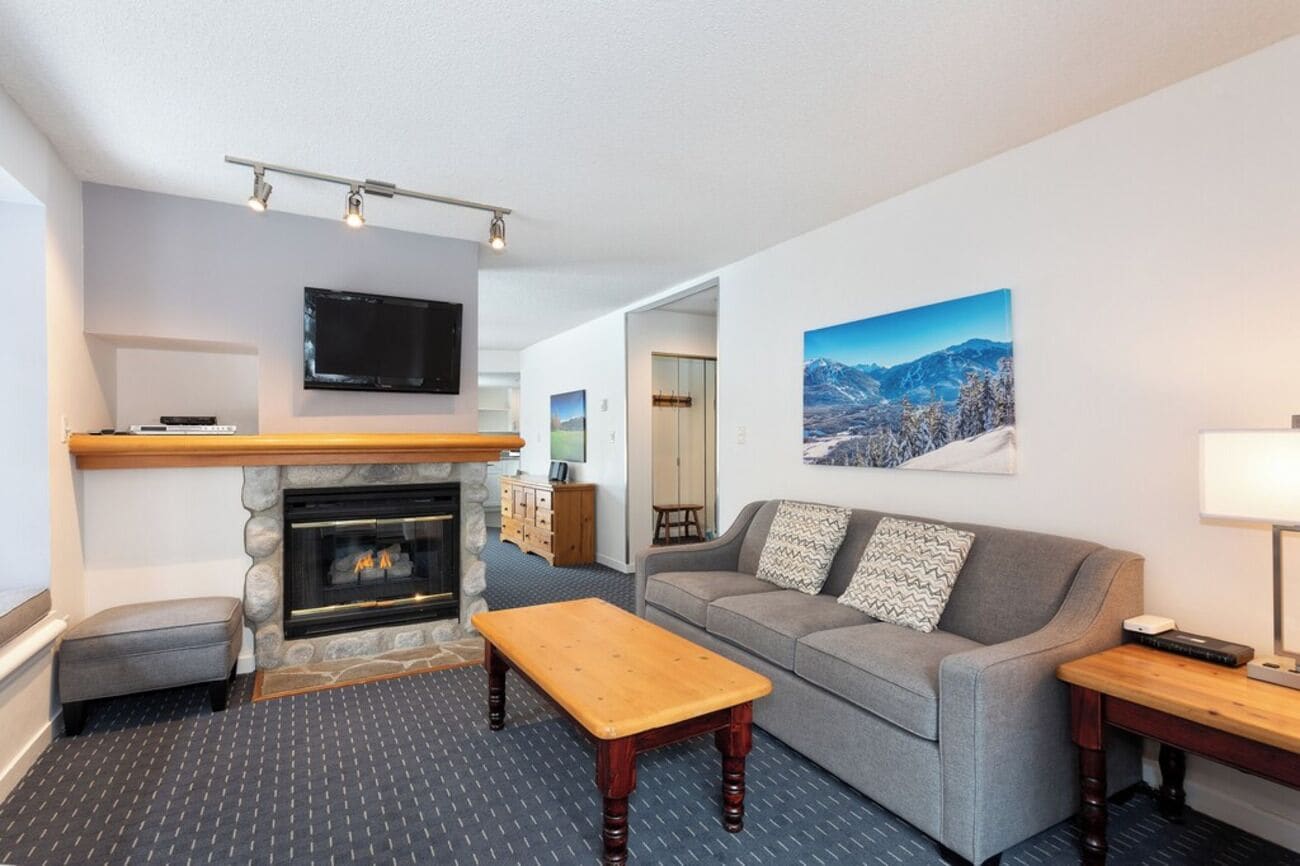 Un salon avec une cheminée, un canapé, une table basse, un téléviseur fixé au-dessus de la cheminée et un tableau représentant des montagnes accroché au mur.