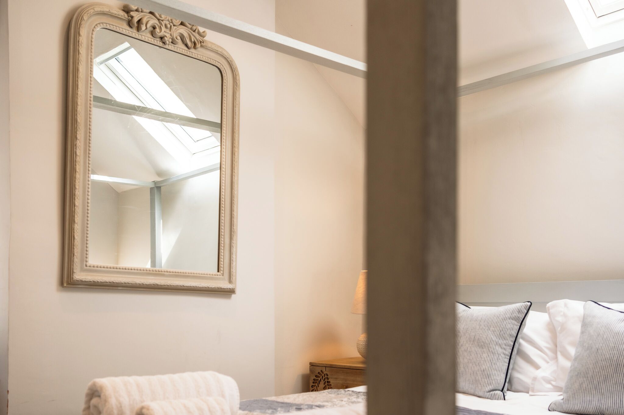 A bedroom with a large mirror, a bed with pillows, a bedside table with a lamp, and a window with a skylight.