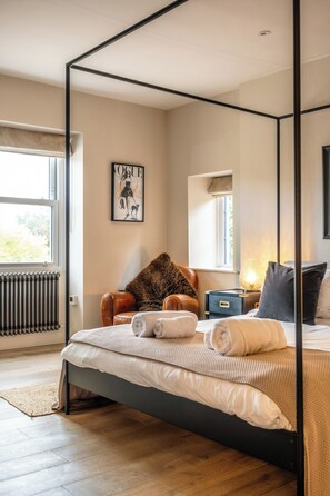Un dormitorio con cama con dosel, un radiador, una ventana con vistas, una silla y una mesita de noche con lámpara.