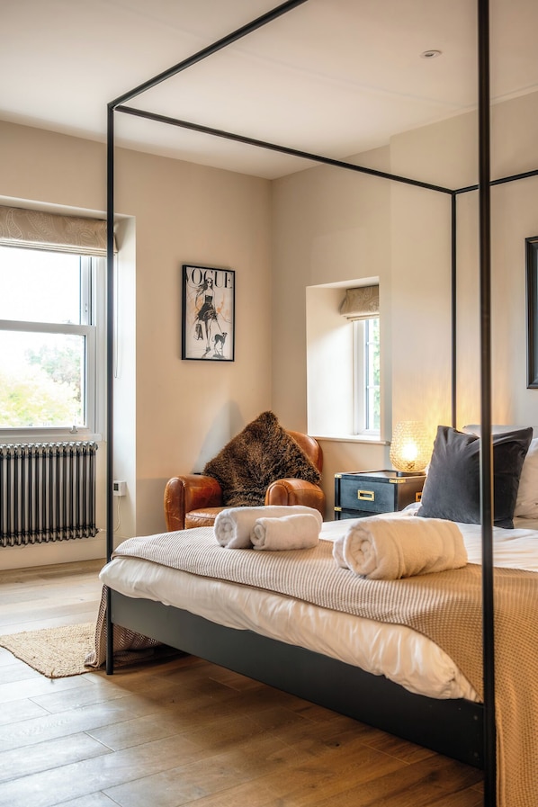 Ein Schlafzimmer mit einem Himmelbett, einem Heizkörper, einem Fenster mit Aussicht, einem Sessel und einem Nachttisch mit Lampe.