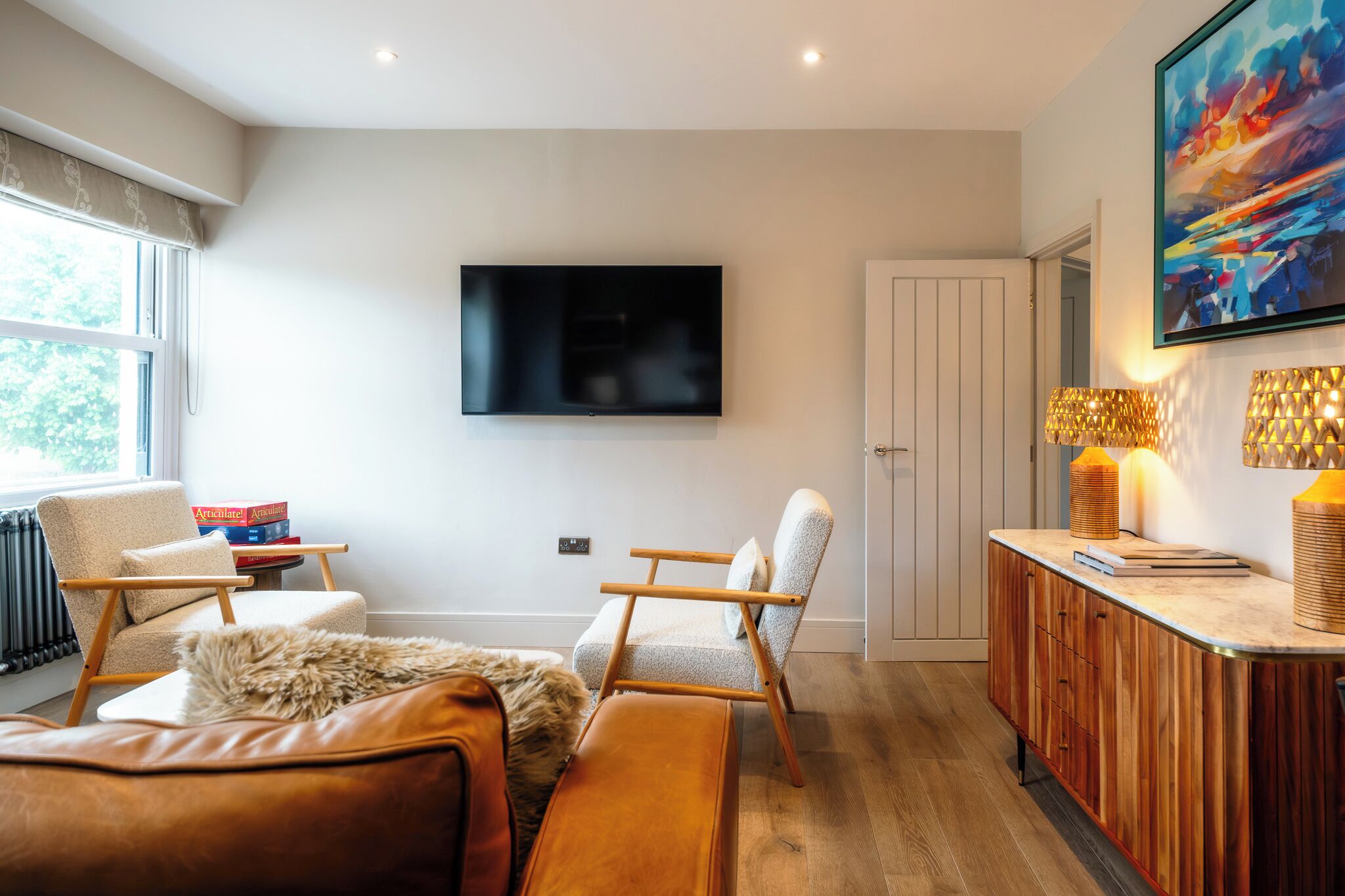Een moderne woonkamer met een flatscreen televisie, twee fauteuils, een bank, een houten kast en een schilderij aan de muur.