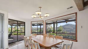 Un espace repas avec une grande table en bois et des chaises blanches, de grandes fenêtres offrant une vue sur un paysage montagneux, et un lustre moderne.