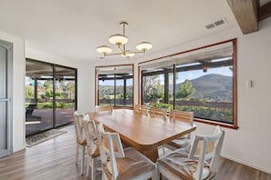 Uma sala de jantar com uma grande mesa de madeira e cadeiras brancas, janelas amplas com vista para uma paisagem montanhosa e um lustre moderno.