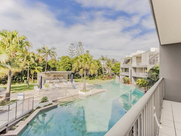 Piscina com espreguiçadeiras, palmeiras e um prédio com varandas.