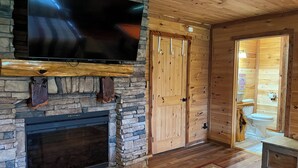 Una chimenea de piedra con un televisor de pantalla plana encima, paredes de madera, una puerta de madera y un baño pequeño visible a través de una puerta abierta.