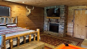 Una habitación tipo cabaña de troncos con una cama, una chimenea de piedra, una cabeza de venado montada y un televisor.