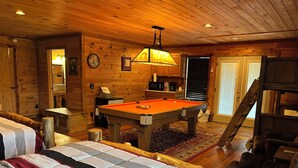 Una habitación acogedora con una mesa de billar, paredes de madera, una cama con manta a rayas y una pequeña cocina.