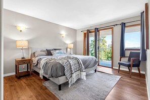 A bedroom with a bed, two bedside lamps, a chair, a window with curtains, and a balcony. - Cozy 3BD Retreat, North Van Balcony Views (North Vancouver)