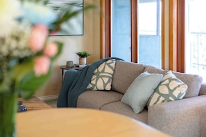 A living room with a beige sofa, a small table with a plant, and a window with curtains. - Cozy 3BD Retreat, North Van Balcony Views (North Vancouver)