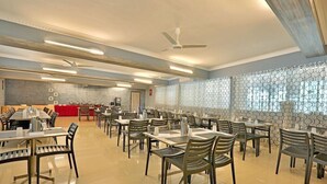 A spacious dining area with multiple tables and chairs, ceiling fans, and a patterned wall. - Corporate Stays (Chengalpattu)