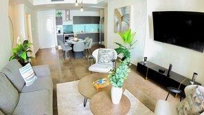 Ein modernes Wohnzimmer mit Sofa, Couchtisch und Fernseher. Im Hintergrund befindet sich ein Essbereich mit Stühlen und eine Küche.