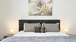 Una camera da letto con un letto, due lampade da comodino, una testiera grigia e un quadro astratto appeso alla parete.