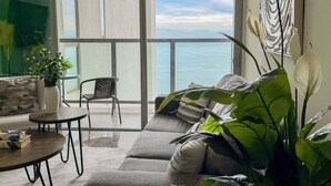 Un salon moderne avec un canapé, une petite table et un fauteuil, un balcon avec vue sur la mer et une grande plante.