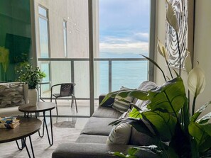 Ein modernes Wohnzimmer mit einer Couch, einem kleinen Tisch und einem Sessel, ein Balkon mit Meerblick und eine große Pflanze.