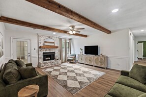 A living room with a fireplace, wooden beams, a ceiling fan, a television, and a sofa. - 5BR Near Graceland Sleeps 10 with Piano, Fire Pit, and Games (Memphis)
