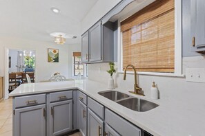 A kitchen with grey cabinets, a white countertop, a stainless steel sink, and a window with blinds. - 5BR Near Graceland Sleeps 10 with Piano, Fire Pit, and Games (Memphis)