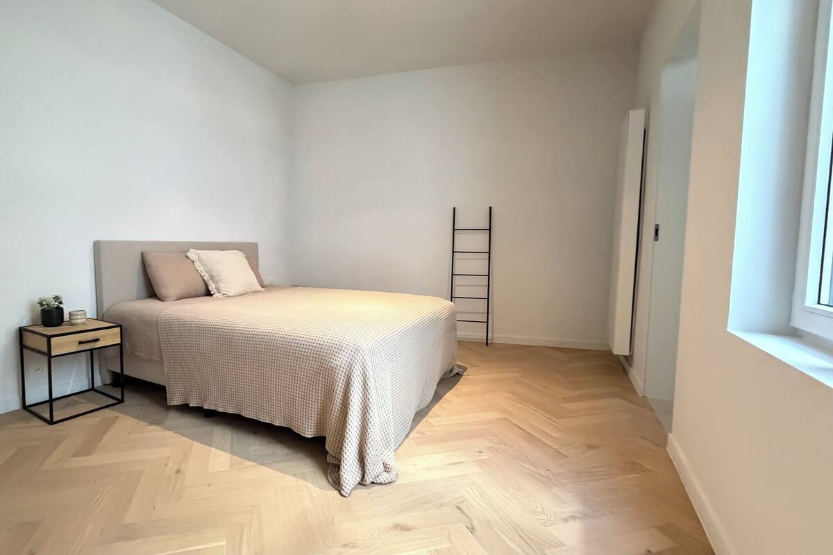Quarto com cama, mesa de cabeceira e escada encostada à parede.