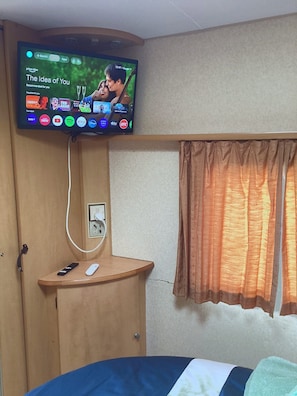 Habitación de hotel con televisor montado en la pared, una mesita de noche con control remoto, toma de corriente y ventana con cortinas.
