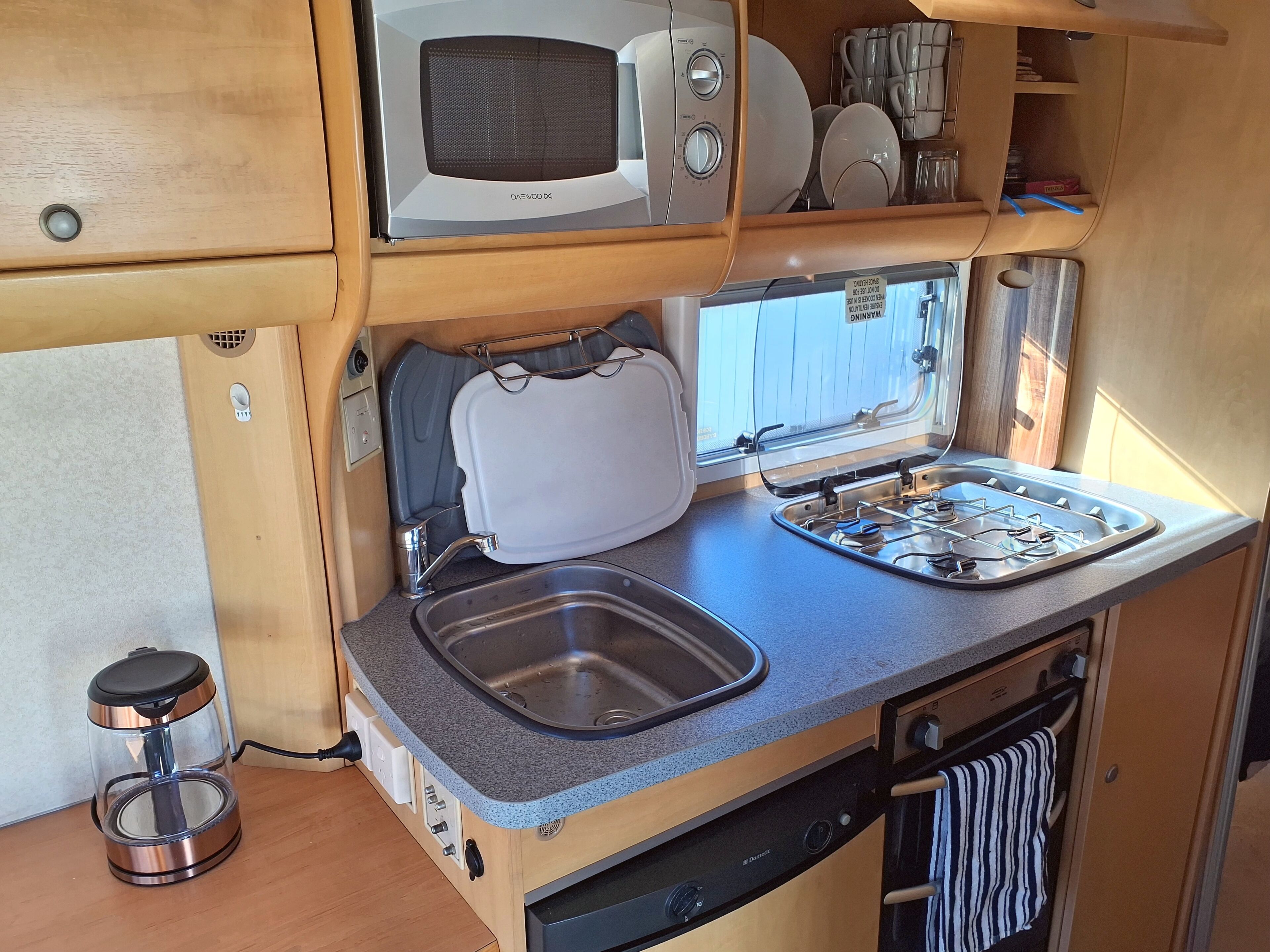 A compact kitchenette with a microwave, sink, stove, and storage shelves.