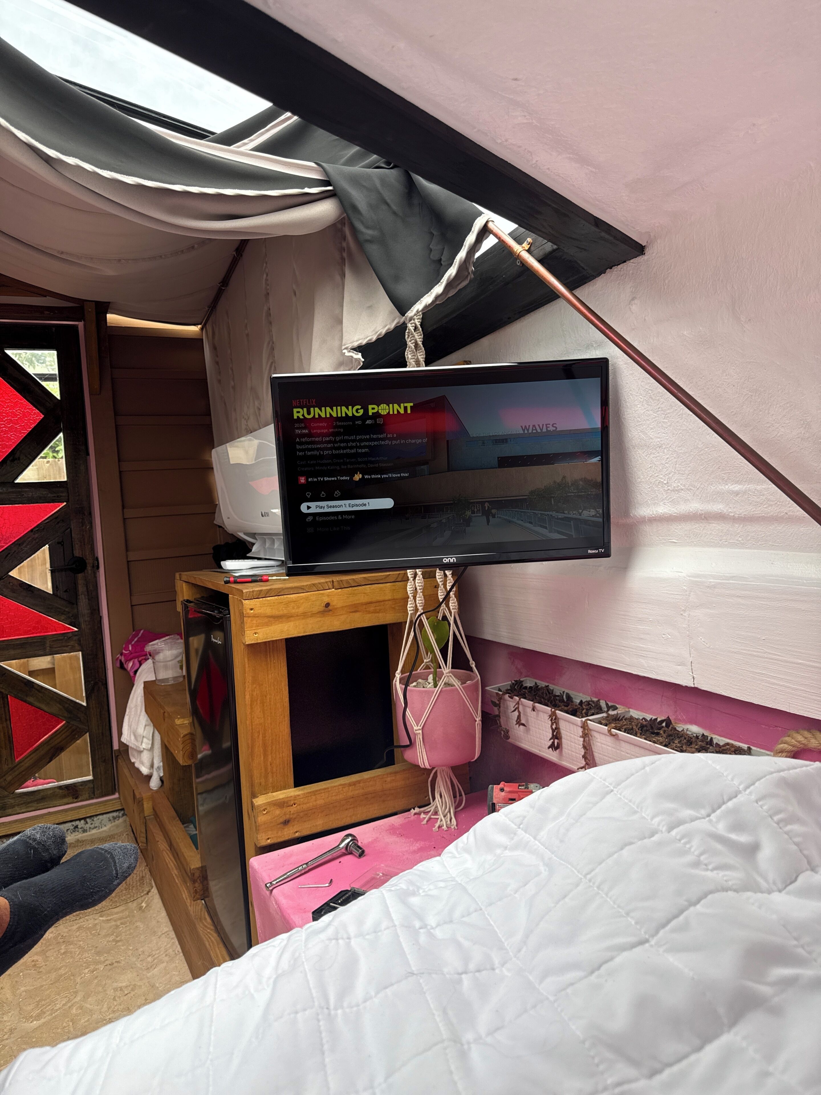 Een slaapkamer met een bed, een aan de muur bevestigde televisie, een houten kast, een roze plant en een deur met een geometrisch ontwerp.