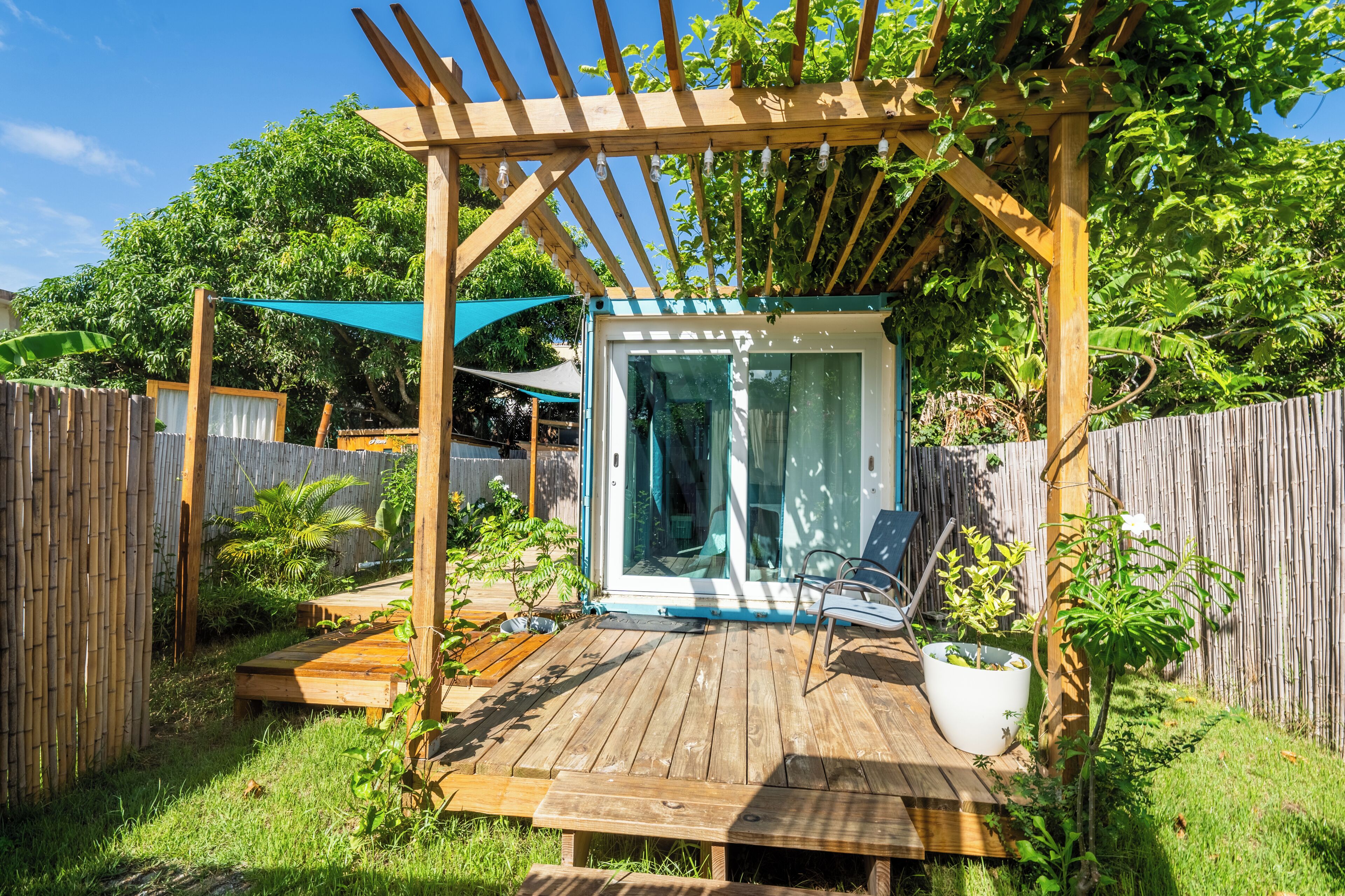 A wooden deck with a glass door leading to a covered area, surrounded by a bamboo fence and lush greenery.