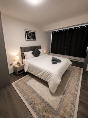 Et moderne soveværelse med en stor seng, et sengebord med lampe, et indrammet billede på væggen og et vindue med gardiner.