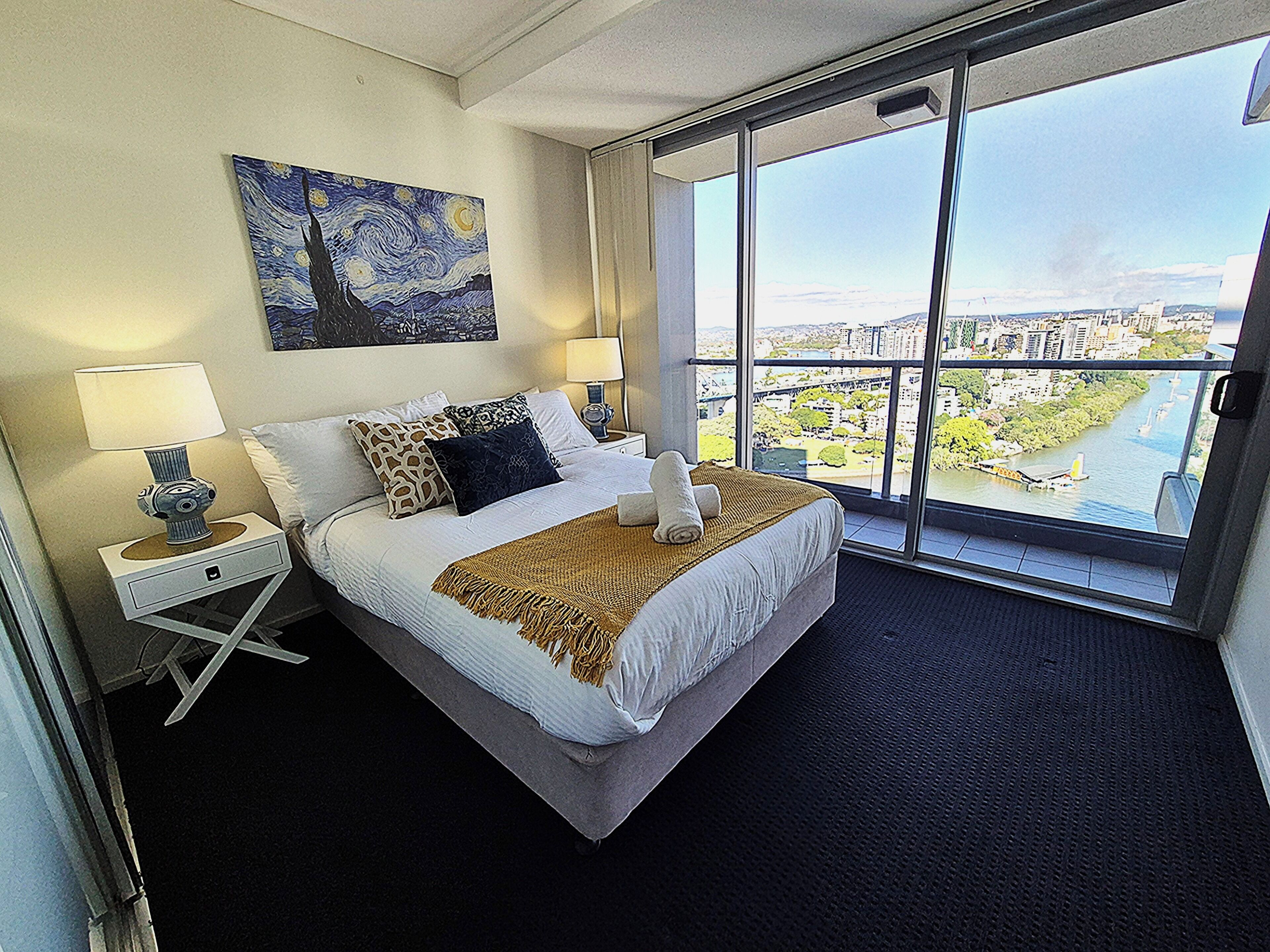 A hotel room with a large bed, two bedside tables, a nightstand, a painting on the wall, and a view of a cityscape through a large window.