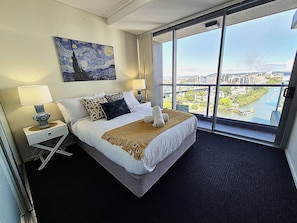 Habitación de hotel con una cama grande, dos mesitas de noche, un atril, un cuadro en la pared y vistas de un paisaje urbano a través de un ventanal amplio.