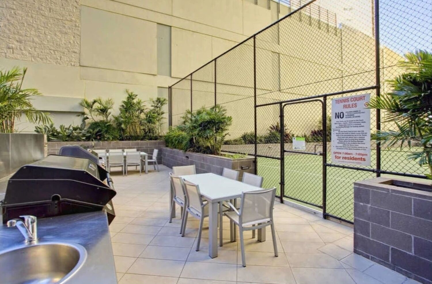 Een terras buiten met een barbecue, een zithoek en een tennisbaan zichtbaar door een hek.