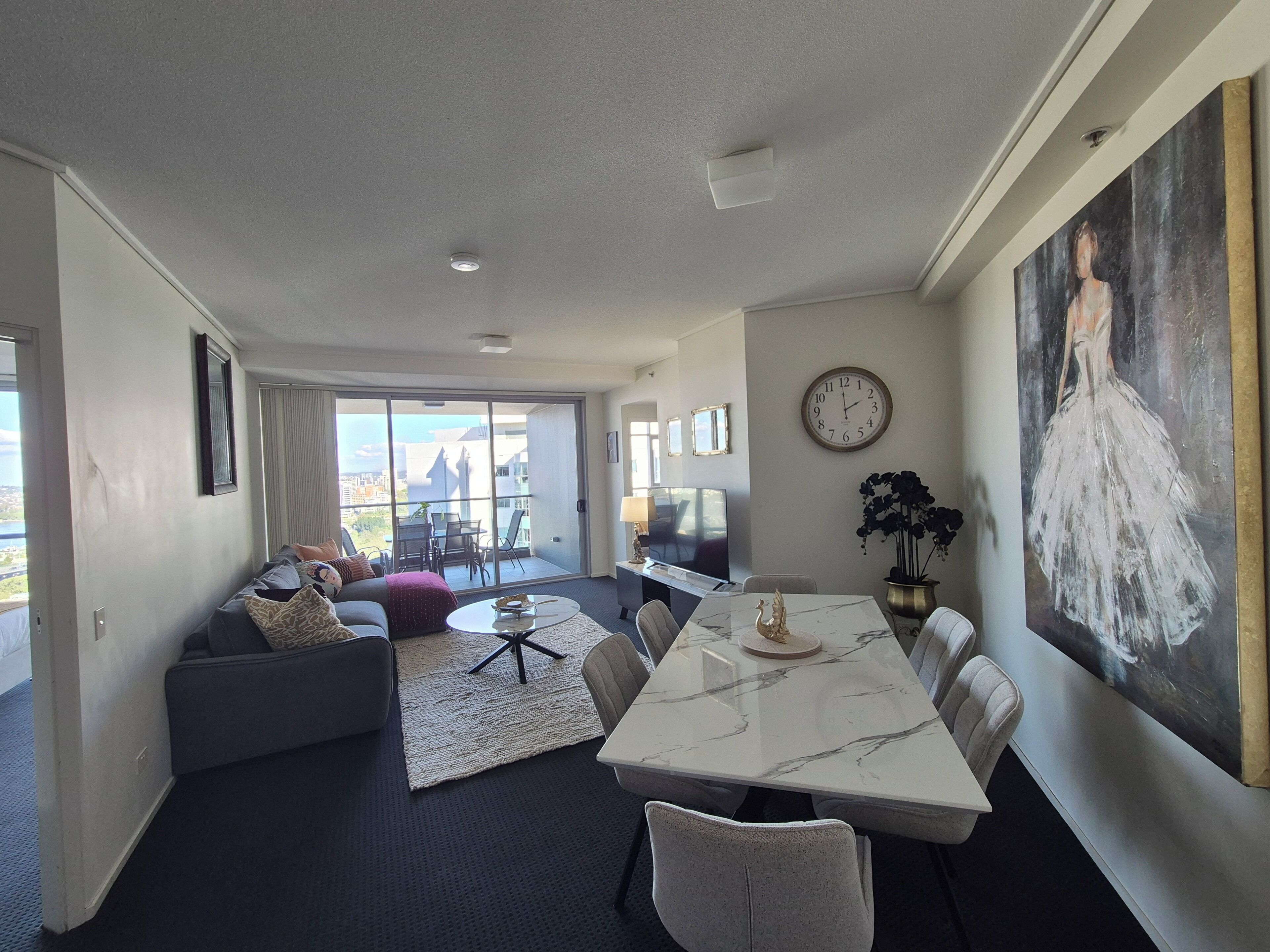 A modern living room with a large window, a dining area with a marble table, and a painting on the wall.