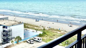 Uitzicht op een strand vanaf een balkon, met een zwembad, een gebouw en een geparkeerde auto.