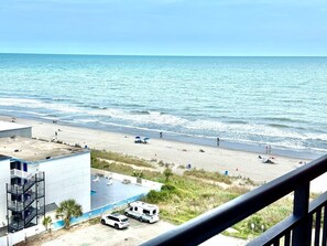 Blick auf einen Strand vom Balkon aus, mit Pool, Gebäude und geparktem Fahrzeug.