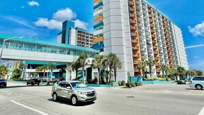 Een meerhoog hotel met een modern ontwerp, met een opvallende ingang met een blauw dakzeil, een parkeerplaats en een helderblauwe lucht.