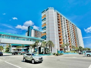 Ein mehrstöckiges Hotel mit modernem Design, das einen markanten Eingangsbereich mit einem blauen Vordach, einen Parkplatz und einen strahlend blauen Himmel zeigt.