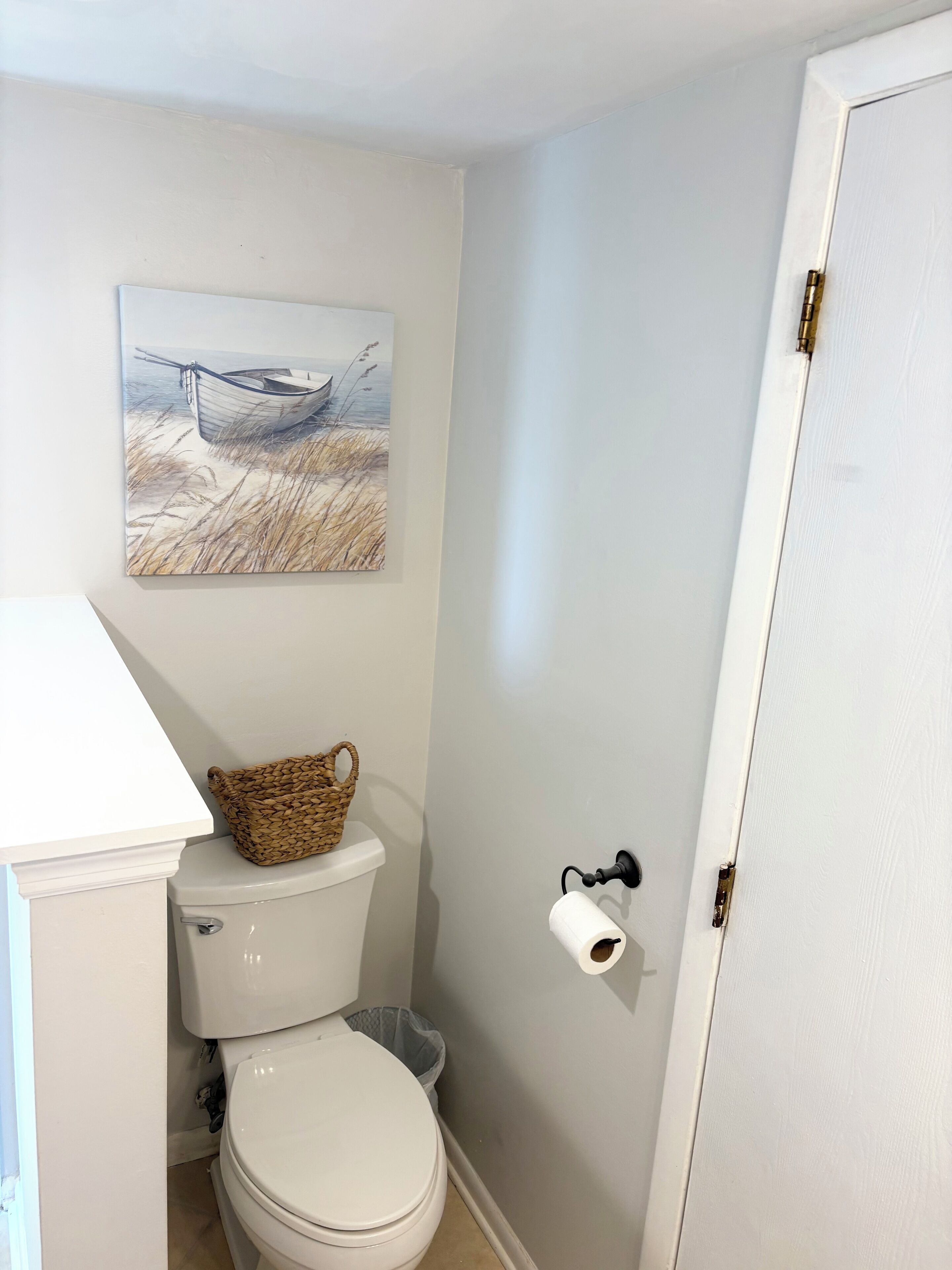 Un baño con inodoro, una cesta sobre el tanque, un rollo de papel higiénico y un cuadro en la pared.