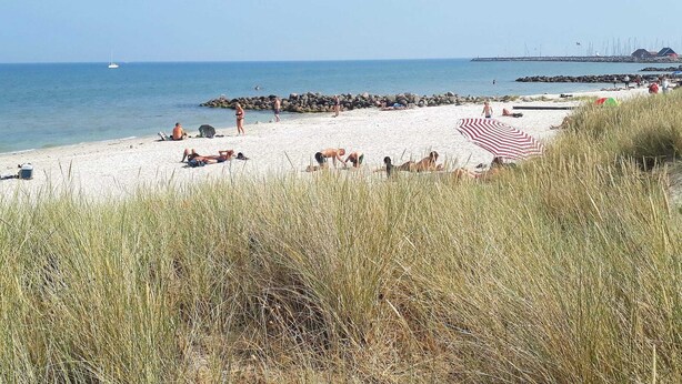 A beach with people sunbathing, a large red and white striped umbrella, and a rocky breakwater in the background. - 6 Person Holiday Park Home in Saeby (Sæby)