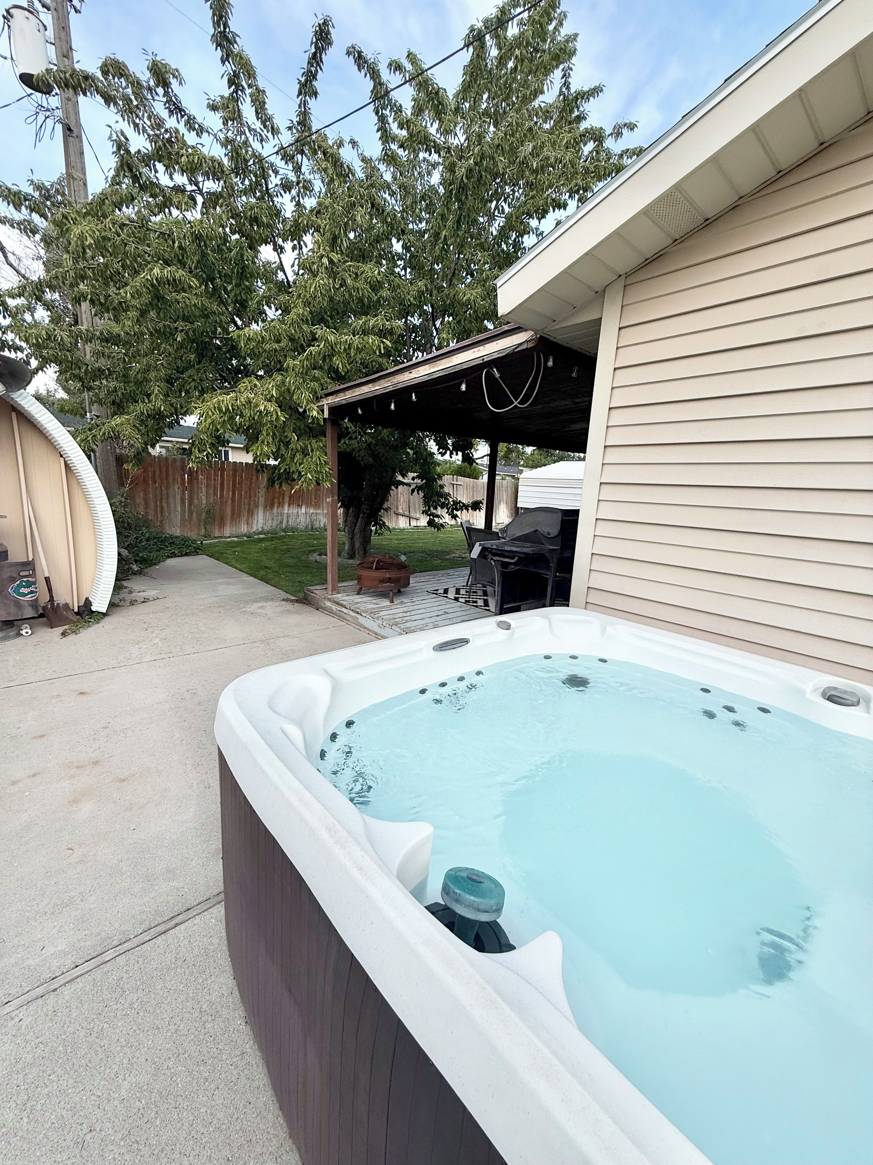 Un jacuzzi en un patio trasero con un porche cubierto y una valla de madera al fondo.